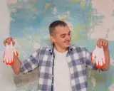 Homme souriant tenant des gants blancs à bouts orange devant un mur texturé. Il semble prêt à peindre, peut-être pour estimer le prix peinture m2 au black.