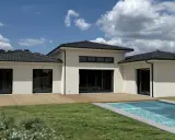 Une maison du sud moderne avec piscine, terrasse en bois et grand jardin sous un ciel bleu nuageux.