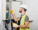 Un électricien artisan, casque blanc et gants jaunes, manipule des fils dans un tableau électrique.