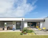 Une jeune personne traverse la pelouse devant une petite maison moderne avec une terrasse aménagée et un jardin de gravier.