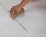 Main d'une personne nettoyant le joint de carrelage avec une éponge. La couleur du joint est sombre, contrastant avec le carrelage clair.