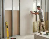 Un homme utilise une perceuse pour poser du placo sur une structure métallique. Des outils et des plaques de plâtre sont visibles.