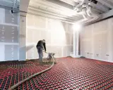 Un ouvrier installe un système de chauffage au sol hydraulique. Des tuyaux rouges serpentent sur une grille noire, prêts à diffuser une chaleur douce.