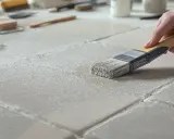 Application d'une couche de béton ciré sur du carrelage. Une main tient un pinceau pour étaler le produit sur les carreaux.
