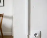 Une goulotte blanche est fixée verticalement sur un mur blanc, dissimulant des câbles. Une prise électrique est visible en bas à droite.