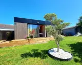 Un charmant pavillon maison moderne avec terrasse en bois, entouré d'un jardin verdoyant et d'un petit olivier.