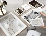 Un mur de cadre couloir prend forme avec des œuvres abstraites et photographiques. Un marteau et des clous sont prêts pour l'accrochage.
