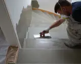 Un ouvrier, masqué et ganté, applique un enduit sur les marches d'un escalier en cours de restauration.