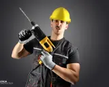 Un homme en casque jaune, gants et tenue de travail tient une perceuse. Il a une expression faciale exagérée, comme un architecte ou un constructeur prêt à relever un défi.