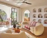 Comment habiller un mur blanc salon : des niches décorées d'objets ethniques et de poteries, un canapé incurvé, des fauteuils bouclette, et une vue sur un jardin luxuriant.