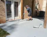Un homme applique une peinture sur mur béton extérieur avec un rouleau. Le sol gris est en cours de rénovation.