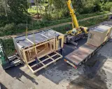 Une grue soulève un module de construction pour des maisons modulaires. Un chariot élévateur et un camion plateau sont également présents.