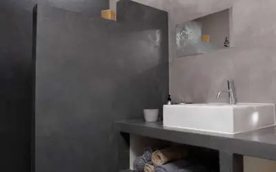 Salle de bain moderne avec murs en béton ciré. Une douche sans joint, un lavabo rectangulaire et des serviettes pliées créent une ambiance épurée.