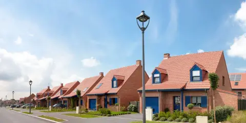 Vue d'une rue bordée de maisons de lotissement aux toits rouges et volets bleus, sous un ciel d'été.