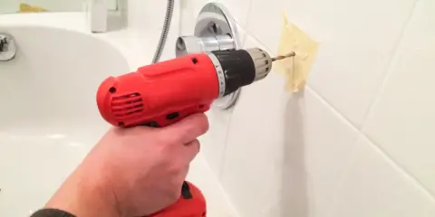 Une main tient une perceuse rouge pour percer du carrelage blanc, avec du ruban adhésif pour guider le foret.