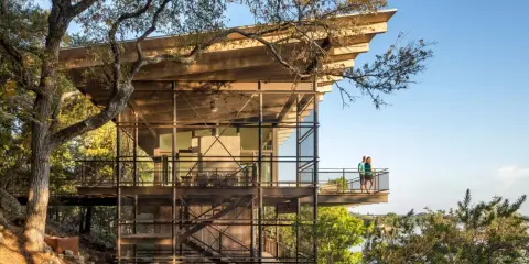 Maison moderne sur un terrain en pente raide, avec des balcons et des escaliers extérieurs en métal, nichée dans les arbres près d'un lac.