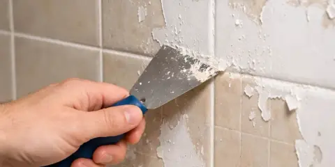 Main tenant enlever peinture sur carrelage avec une spatule. La peinture s'écaille sur les carreaux beiges.
