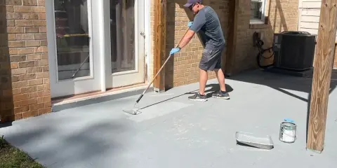 Un homme applique une peinture sur mur béton extérieur avec un rouleau. Le sol gris est en cours de rénovation.