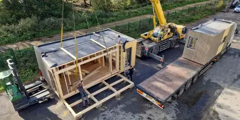 Une grue soulève un module de construction pour des maisons modulaires. Un chariot élévateur et un camion plateau sont également présents.