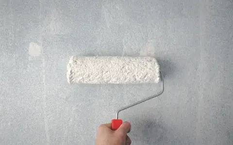Main tenant, une main tient un rouleau à peinture blanc sur un mur bleu clair. Les traces de rouleau à la lumière révèlent la texture du mur.