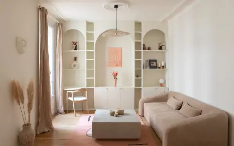 Un coin bureau discret dans un salon cosy, avec une étagère murale arche, un canapé beige et un tapis rose.