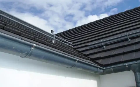 Le chêneau de cette maison, en zinc, est bien fixé sous le toit de tuiles sombres. Le ciel bleu et nuageux promet une belle journée.