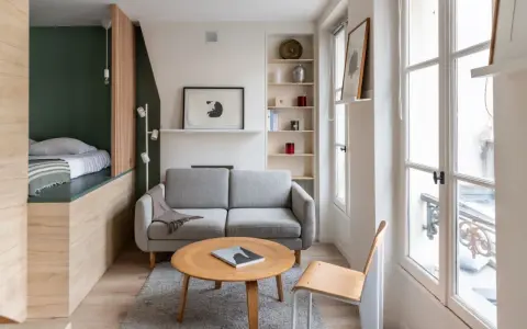 Aménagement petit appartement : lit surélevé, canapé gris, table basse ronde en bois, étagères et grande fenêtre lumineuse.