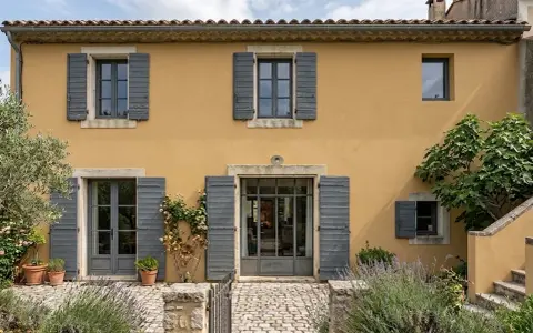 Façade d'une maison provençale à l'enduit projeté ocre, avec volets gris et végétation luxuriante.