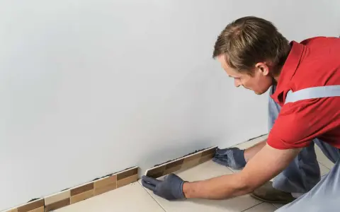 Un homme en tenue de travail enfile des gants pour enlever la plinthe du carrelage.