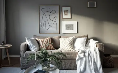 Salon cosy avec un **canapé gris clair** orné de coussins texturés et d'une couverture blanche. Des œuvres d'art abstraites décorent le mur.