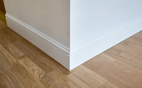Un coin de mur blanc avec des plinthes moulurées et un parquet en bois clair. Ces plinthes servent à protéger les murs et à cacher les imperfections.