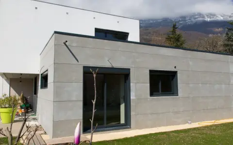 Extension moderne en parpaing gris, 20m2, avec grandes baies vitrées et vue sur montagnes.