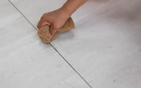 Main d'une personne nettoyant le joint de carrelage avec une éponge. La couleur du joint est sombre, contrastant avec le carrelage clair.