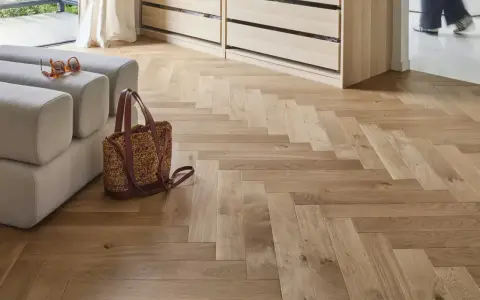 Un parquet en bois clair au calepinage en chevrons, avec un canapé, un sac et des lunettes posés dessus.