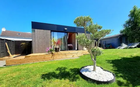 Un charmant pavillon maison moderne avec terrasse en bois, entouré d'un jardin verdoyant et d'un petit olivier.