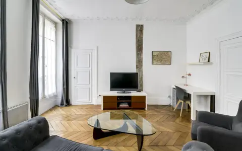 Salon moderne avec canapé gris, table basse en verre, bureau et mur blanc orné d'une carte de Paris.