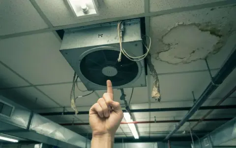Une main pointe un VMC bruyant au plafond, à côté d'une tache d'humidité.