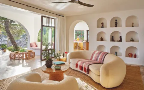 Comment habiller un mur blanc salon : des niches décorées d'objets ethniques et de poteries, un canapé incurvé, des fauteuils bouclette, et une vue sur un jardin luxuriant.