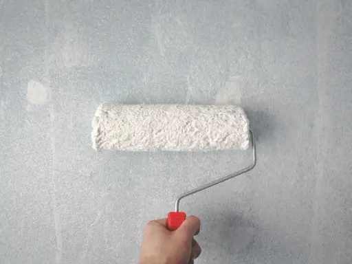 Main tenant, une main tient un rouleau à peinture blanc sur un mur bleu clair. Les traces de rouleau à la lumière révèlent la texture du mur.