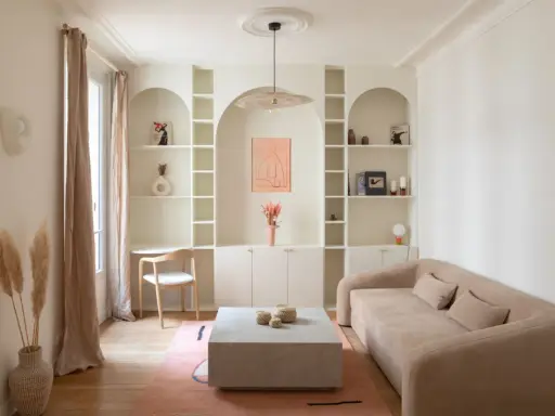 Un coin bureau discret dans un salon cosy, avec une étagère murale arche, un canapé beige et un tapis rose.