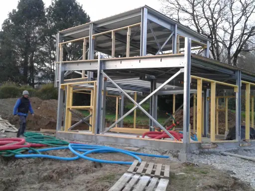 Chantier d'une maison ossature métallique, structure en acier et bois, avec des tuyaux colorés au sol.