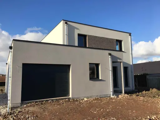 Maison moderne avec garage intégré. La couleur façade maison est un gris clair, contrastant avec des touches de blanc cassé et un bandeau de briques sombres.