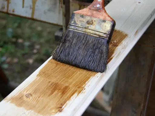 Application d'une lasure bois pour un avant-après visible. Le pinceau dépose une teinte chaude sur le bois brut.