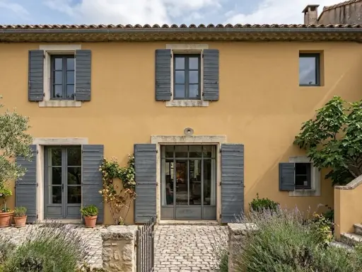 Façade d'une maison provençale à l'enduit projeté ocre, avec volets gris et végétation luxuriante.