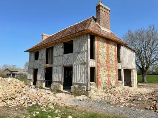 Construction d'une maison normande traditionnelle, avec colombages apparents et murs en pierre et brique. Le chantier est en cours.
