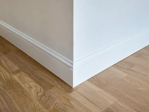Un coin de mur blanc avec des plinthes moulurées et un parquet en bois clair. Ces plinthes servent à protéger les murs et à cacher les imperfections.