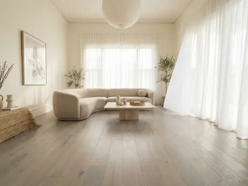 Un salon lumineux avec un canapé incurvé beige, une table basse en pierre et un sol en bois gris clair. Le beige et le blanc s'accordent parfaitement avec le gris clair.