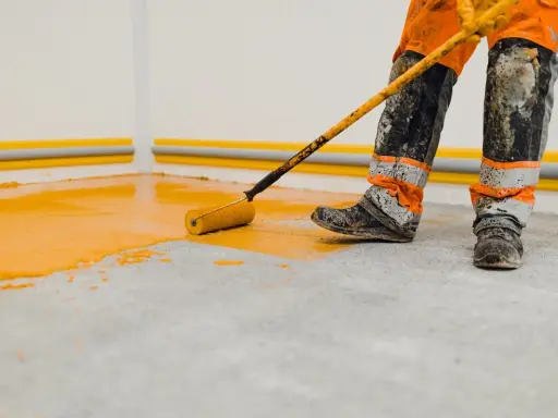 Un ouvrier en combinaison orange et grise, les bottes sales, utilise un rouleau pour peindre le sol d'un garage en jaune vif.