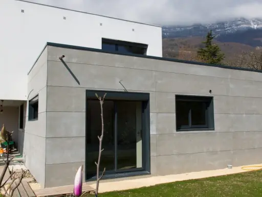 Extension moderne en parpaing gris, 20m2, avec grandes baies vitrées et vue sur montagnes.