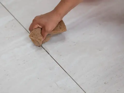 Main d'une personne nettoyant le joint de carrelage avec une éponge. La couleur du joint est sombre, contrastant avec le carrelage clair.